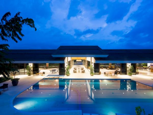 A luxurious building with a large illuminated pool at night, surrounded by seating areas and a clear blue sky overhead.