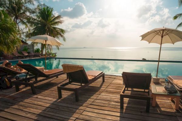 A serene poolside setting with lounge chairs, umbrellas, and palm trees overlooking a tranquil body of water under a bright sky.