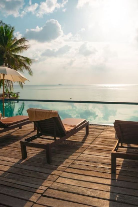 A serene poolside deck with loungers, umbrellas, palm trees, and a view of the ocean under a cloudy sky and bright sun.