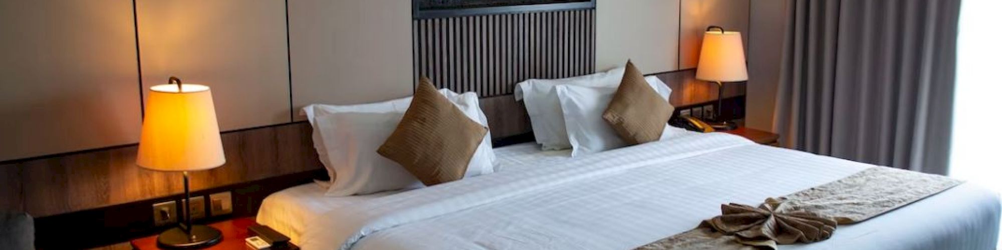 A well-decorated hotel room with a large bed, wall art, bedside lamps, and a window with drapes on the right side.