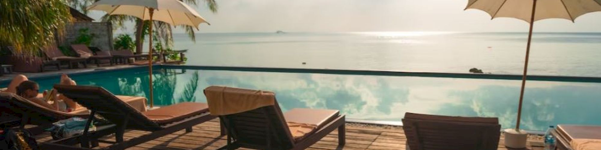 Poolside scene with loungers, umbrellas, and palm trees overlooking a calm sea under a partly cloudy sky.