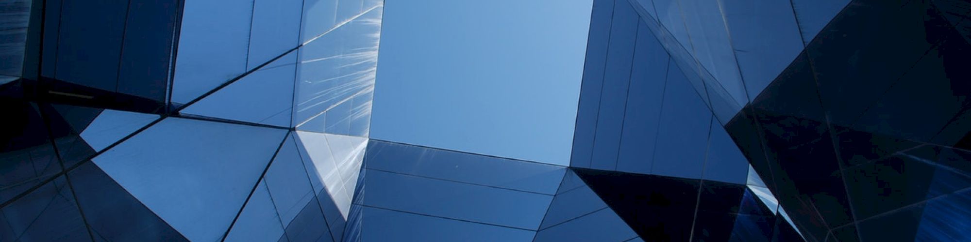 Looking up through a geometric structure of reflective glass panels, showing a clear blue sky framed by angular lines.