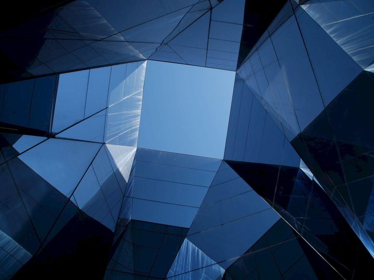 The image shows an upward view of modern reflective glass buildings forming a geometric pattern around a clear blue sky.