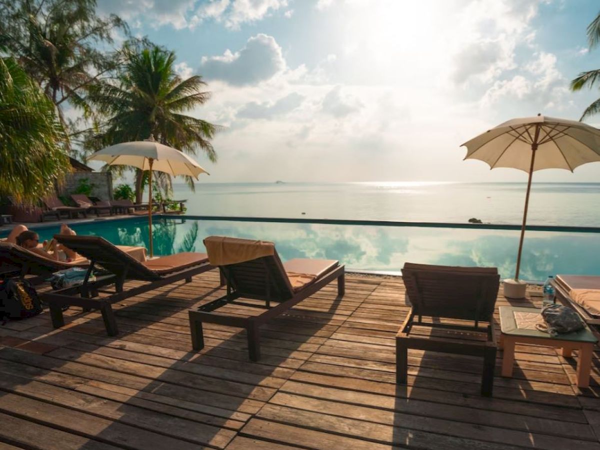 A serene poolside scene with lounge chairs, umbrellas, palm trees, and a view of the sunlit sea and sky on a wooden deck.