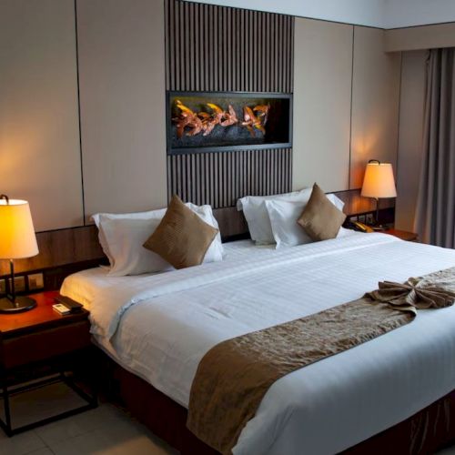 A stylish hotel room with a large bed, bedside tables, lamps, and modern decor, featuring a wall-mounted fireplace.