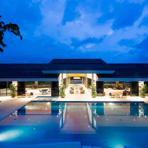 Illuminated building with open architecture, viewed across a large reflective pool under a twilight sky, creating a serene ambiance.