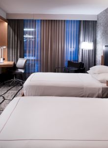 The image shows a modern hotel room with two beds, a wall-mounted TV, a desk, chair, textured wallpaper, and a window with curtains.