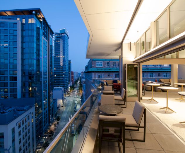 A modern rooftop balcony at dusk, overlooking a city street lined with tall buildings, featuring outdoor seating and tables.