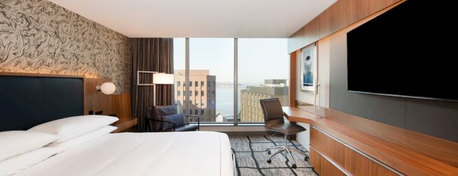 A modern hotel room with a large bed, desk, TV, and city view from the window.