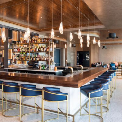 A modern bar with pendant lights, a stocked shelf, blue barstools, and a seating area in the background, creating a cozy atmosphere.