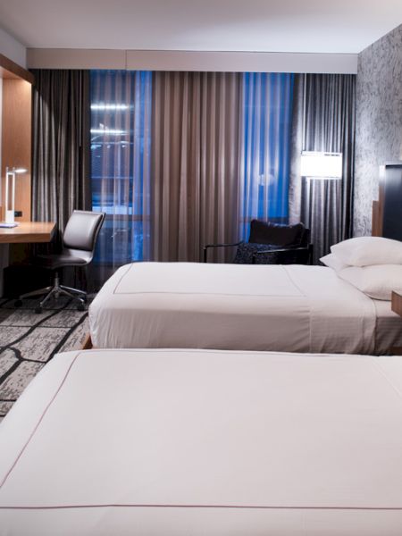 The image shows a modern hotel room with two beds, a desk, large curtains, a TV, and stylish lighting and decor.