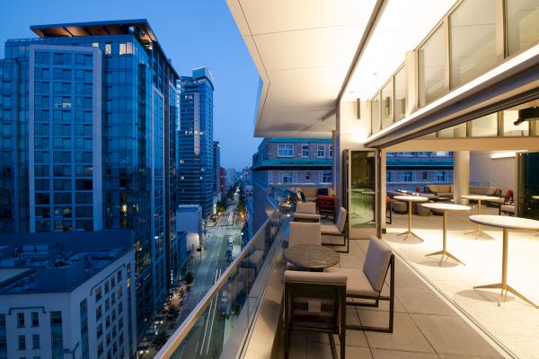 A modern rooftop terrace with seating overlooks a cityscape at dusk. Illuminated buildings line the street, creating a vibrant urban scene.