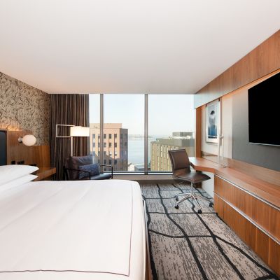 A modern hotel room with a large bed, wall-mounted TV, desk, and city view through wide windows.