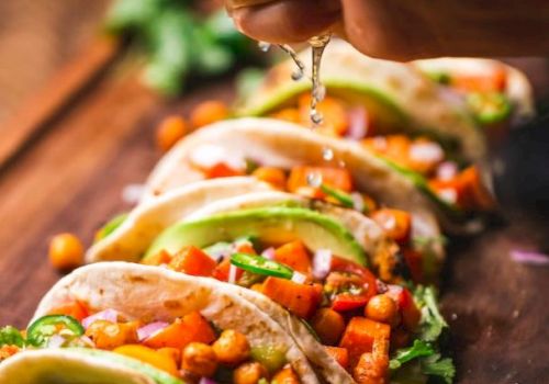 A hand squeezes lime over a row of tacos filled with chickpeas, peppers, avocados, and cilantro on a wooden surface.