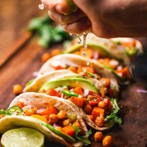 A hand squeezes lime over a row of tacos filled with chickpeas, peppers, avocados, and cilantro on a wooden surface.