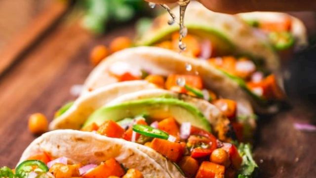 A hand squeezes lime over a row of tacos filled with chickpeas, peppers, avocados, and cilantro on a wooden surface.