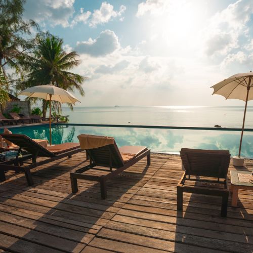 A serene poolside with deck chairs, umbrellas, palm trees, and a calm sea under a bright sky.