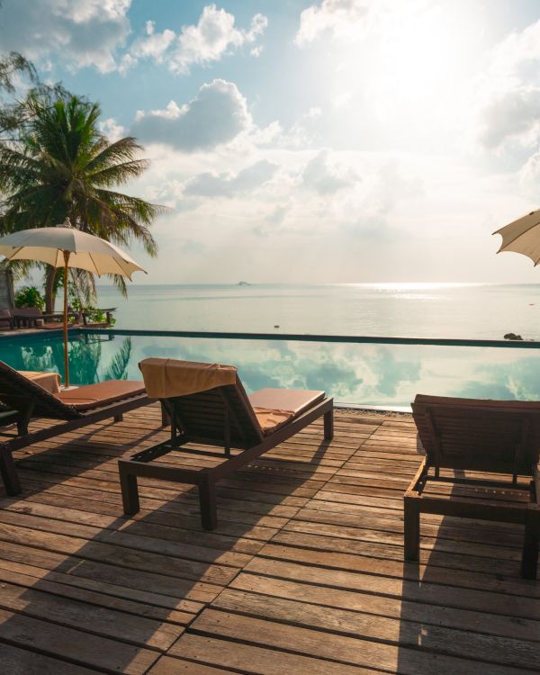 A serene poolside with deck chairs, umbrellas, palm trees, and a calm sea under a bright sky.