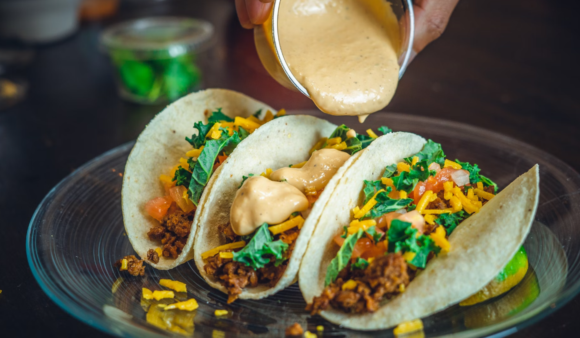 Three tacos filled with ingredients like lettuce, tomatoes, meat, and cheese are being topped with a creamy sauce on a glass plate.