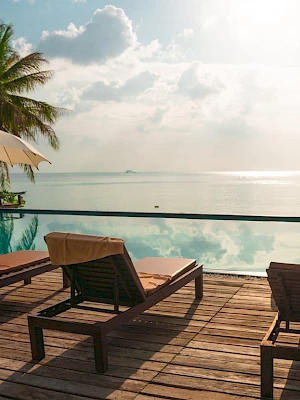 Lounge chairs and umbrellas by an infinity pool overlook the ocean, with palm trees and a cloudy sky enhancing the tranquil scene.