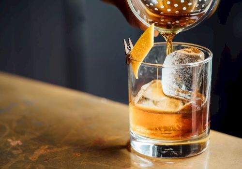 A cocktail being poured into a glass with ice and a citrus peel garnish, set on a wooden surface.