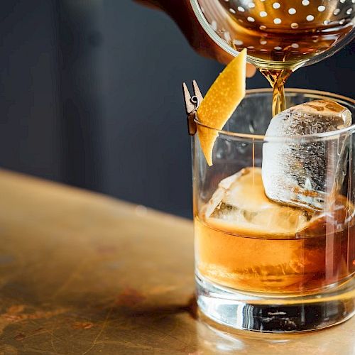 A cocktail being poured into a glass with ice and a citrus peel garnish, set on a wooden surface.