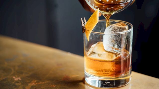 A cocktail being poured into a glass with ice and a citrus peel garnish, set on a wooden surface.