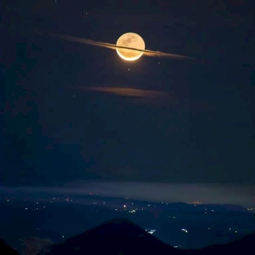 The image shows the moon with clouds resembling Saturn's rings, above a hilly landscape with scattered lights in the distance.
