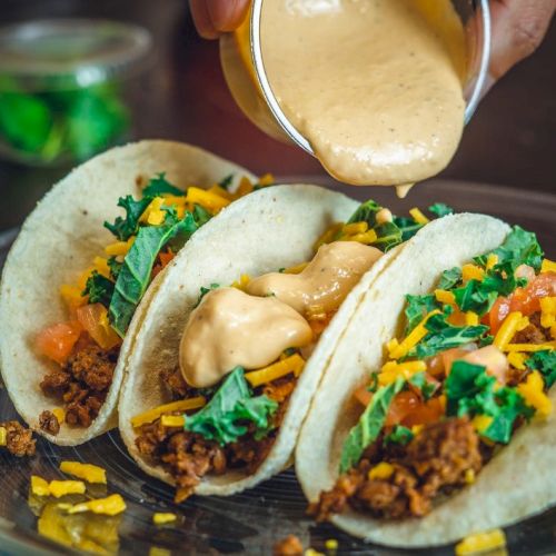 Three tacos filled with meat, cheese, and greens are being topped with a creamy sauce on a plate.