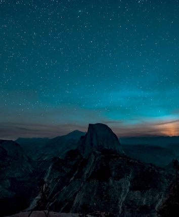 A mountain silhouette under a starry night sky with a glowing horizon and trees in the foreground.