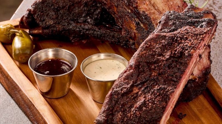 A wooden board with two large pieces of cooked meat, sauce cups, and garnish.