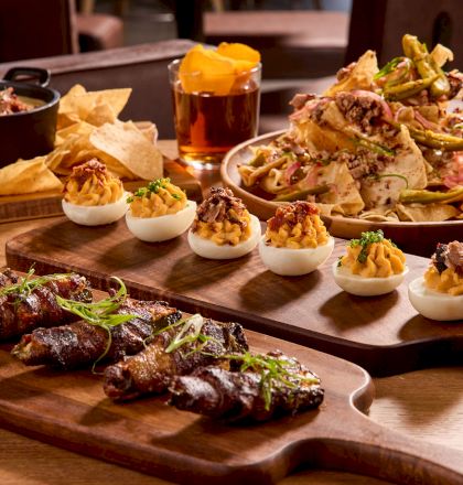 A spread of appetizers includes deviled eggs, bacon-wrapped bites, nachos, chips, and cocktails on wooden boards on a table.