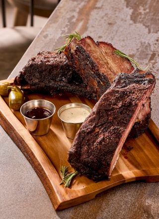 A wooden platter holds grilled beef ribs with two metal sauce cups and garnishes, placed on a textured surface.