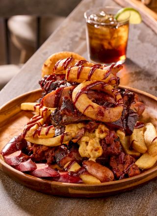 A plate with a mix of fries, sausage, ribs, and onion rings, topped with sauce; a drink with lemon in the background.