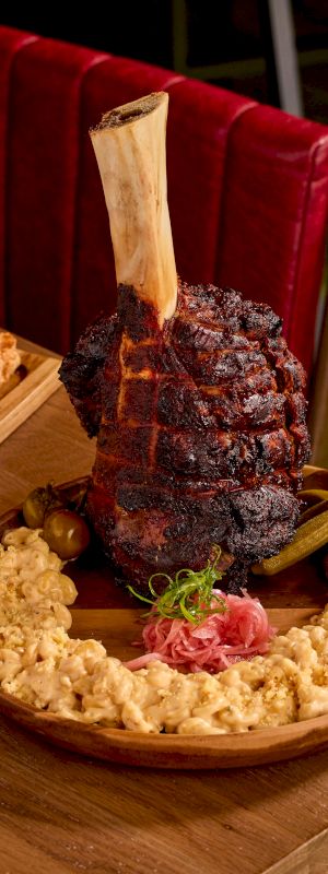 A large, grilled meat piece with a bone on a wooden plate, surrounded by macaroni and garnished with pickles and herbs.