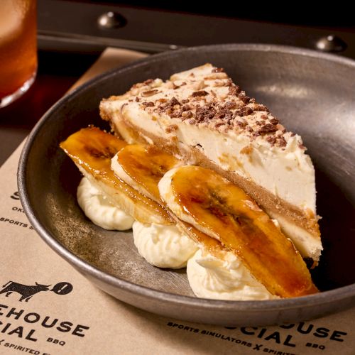 A slice of layered dessert with whipped cream, caramelized banana slices, and chocolate shavings on top, plated on a dark dish.