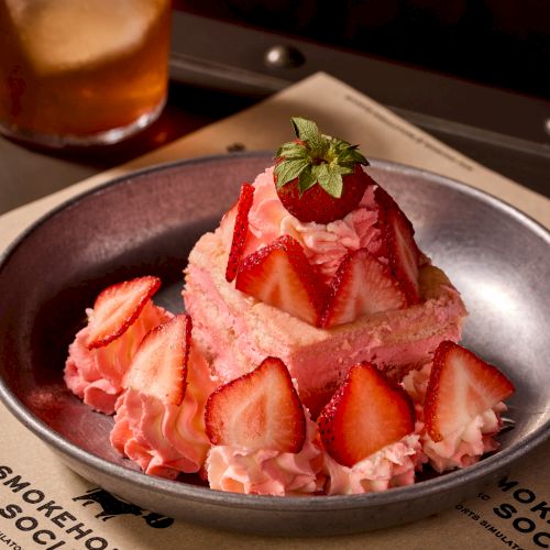 A strawberry-topped pink parfait dessert with layered whipped cream and fresh strawberry slices, served on a dark plate. Delicious treat.
