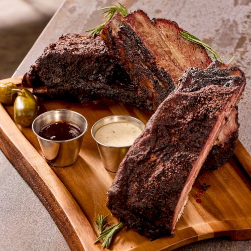 Charred focaccia-style ribs on a wooden board, with two dipping sauces and sprigs of rosemary for garnish. Bon app&eacute;tit.
