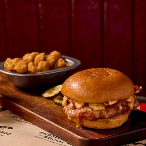 A juicy sandwich with pulled meat on a glossy bun sits on a wooden board, plus a side of crispy fried bites in a bowl; delicious.