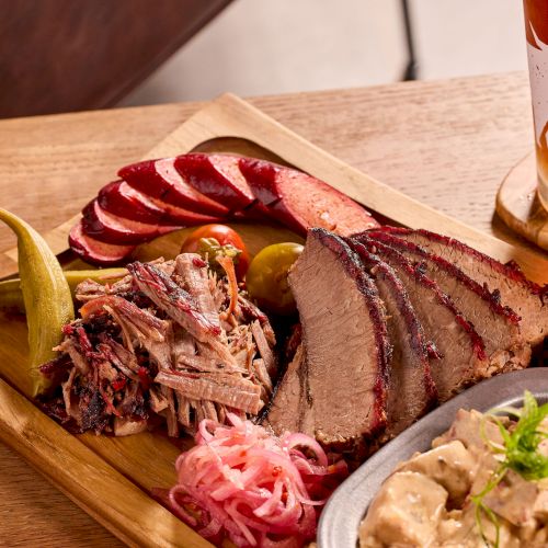 Barbecue platter with brisket, sausage, pickled vegetables, and sides, served with a glass of beer on a wooden table.