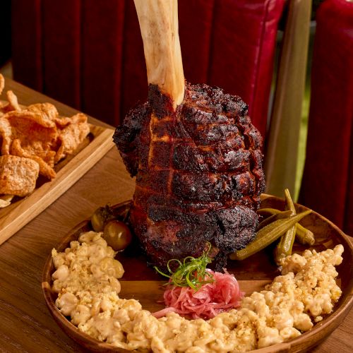 A large roasted meat piece with a bone, on a plate with mac and cheese garnishing, served on a wooden table.