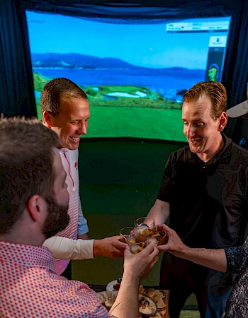 Four people are toasting with drinks in front of a golf simulator displaying a scenic course.