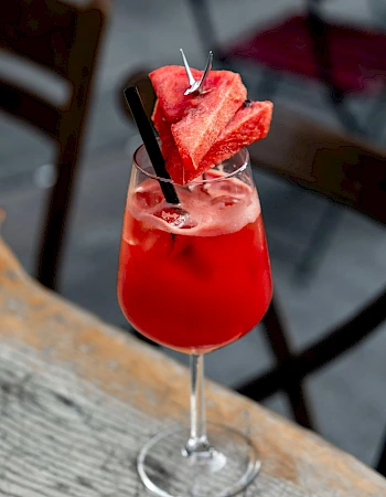 A red cocktail in a glass, garnished with watermelon slices and a cinnamon stick, sits on a wooden table.