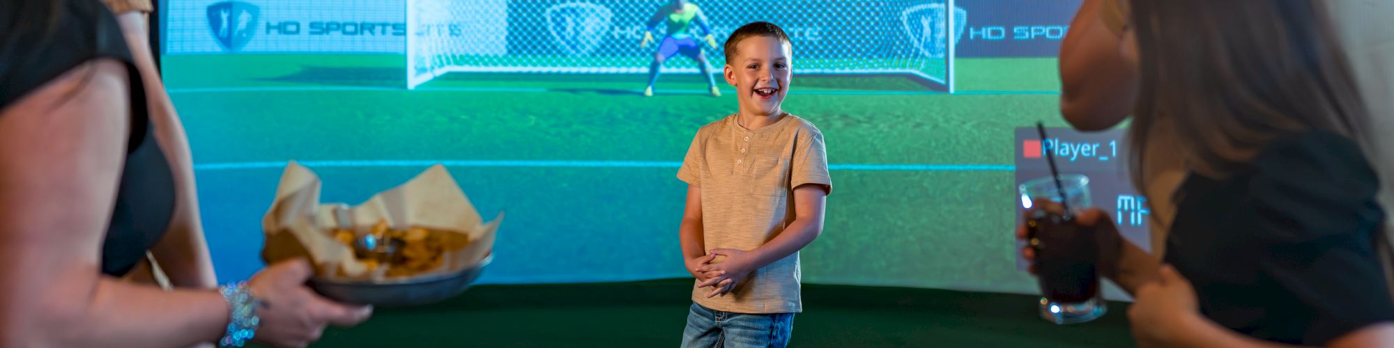 A young boy stands near a soccer ball in front of a digital goal display. Onlookers watch, with refreshments and snacks in hand.