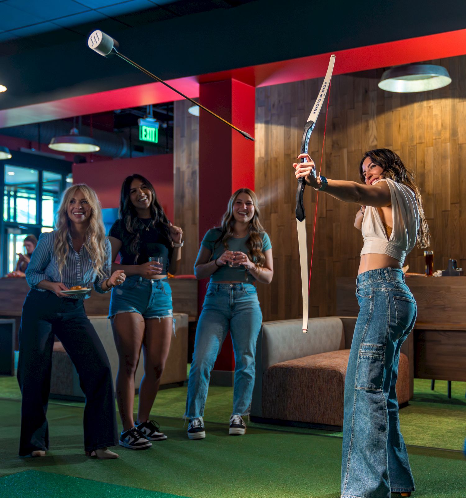 Four people indoors; one is shooting a bow and arrow, others watching and smiling, in a leisure setting.