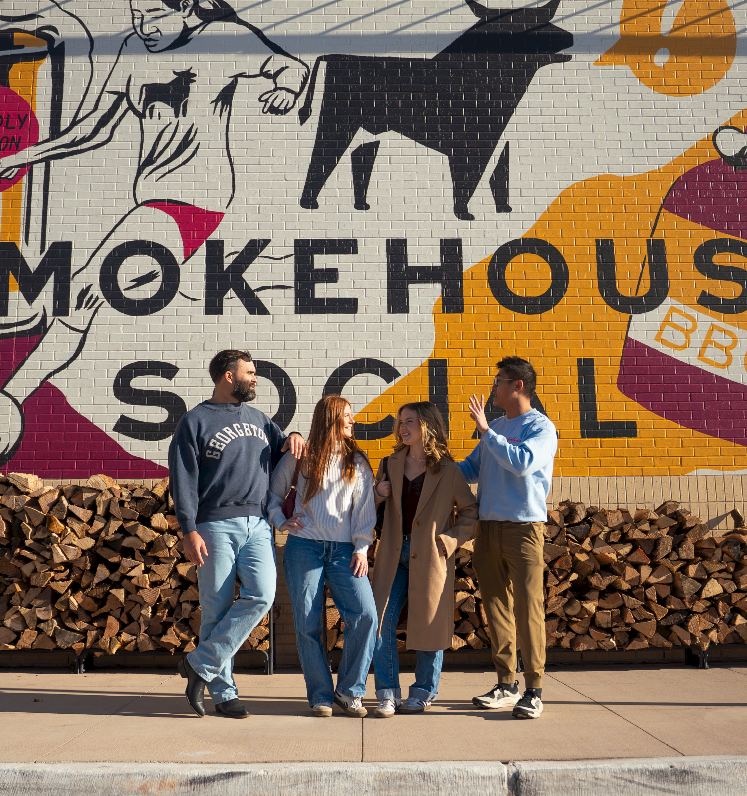 A group of four friends chats in front of a colorful mural reading &ldquo;SMOKEHOUSE&rdquo; with stacked firewood behind them, sunny day, casual vibes.