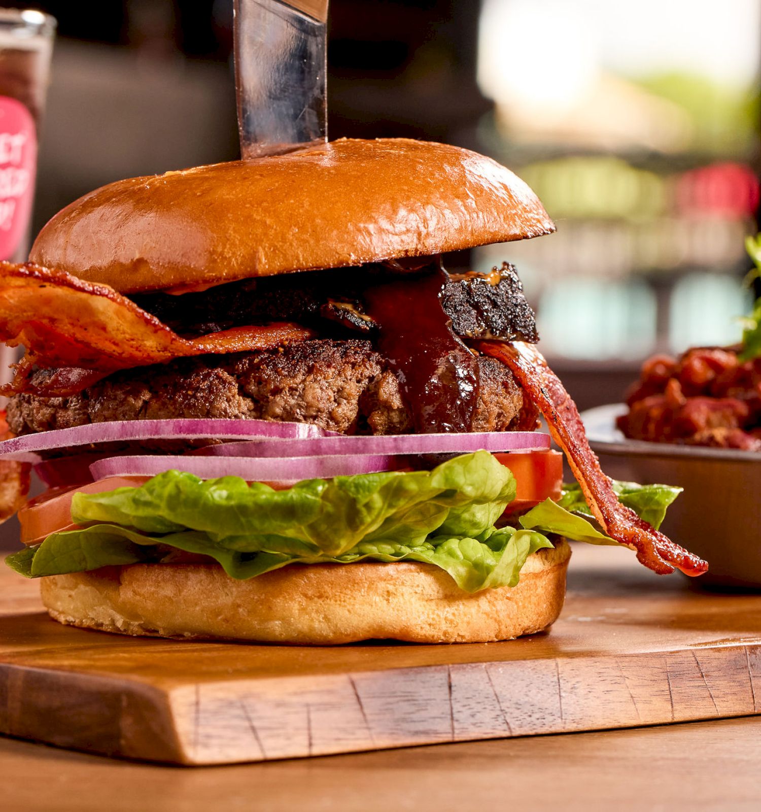 A burger with bacon, lettuce, and onion on a wooden board, accompanied by a bowl of chili and a glass of dark beer in the background.