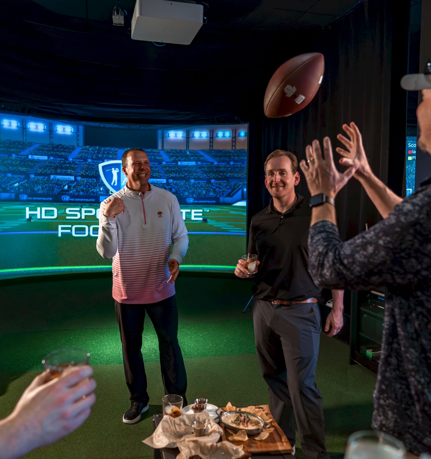 Two men cheer in a sports bar booth while guests watch an HD sports game on a big screen under blue lighting.