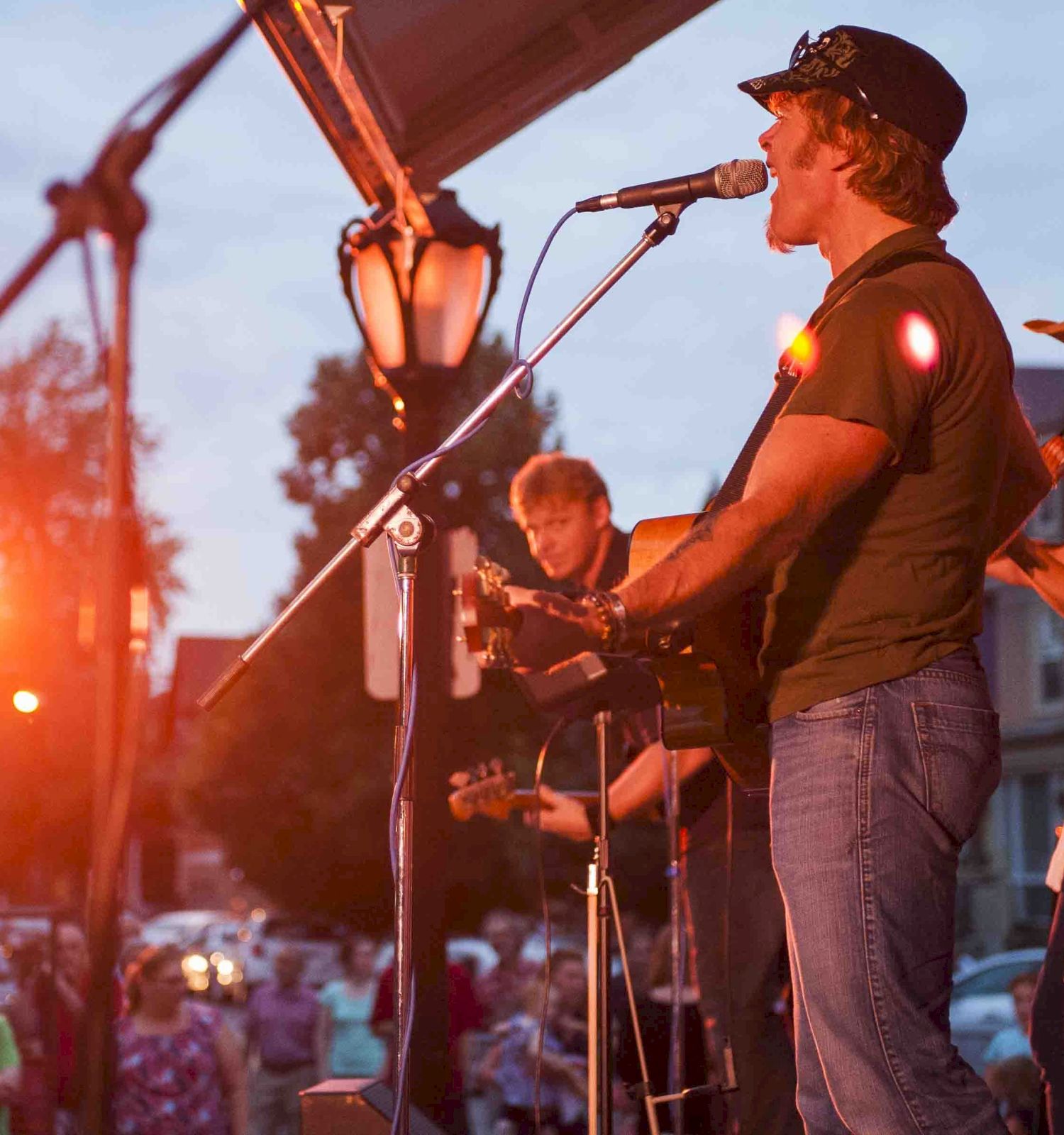 A live outdoor band performs on stage at sunset, guitars and mic in hand, audience watching intently.