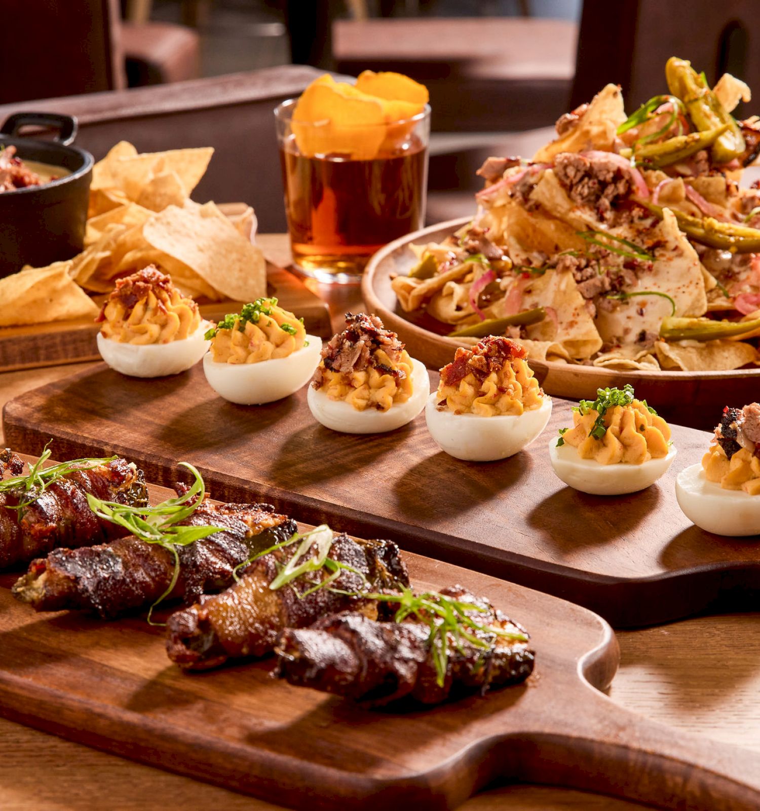 The image shows a variety of appetizers, including nachos, deviled eggs, and bacon-wrapped items, with drinks on a wooden table.